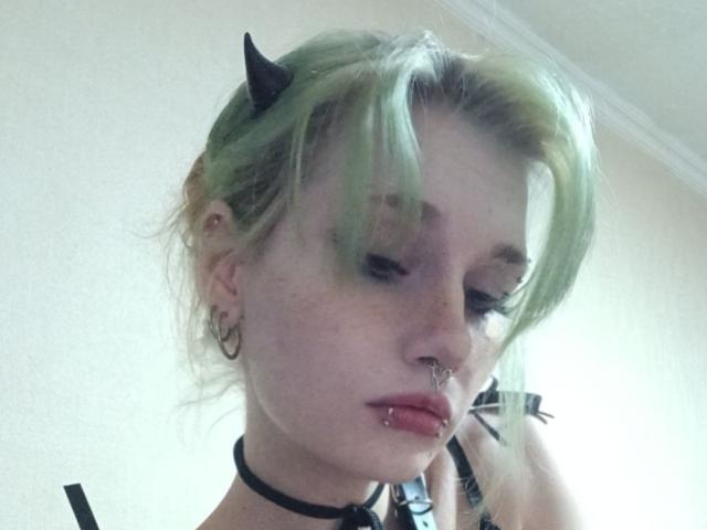 ShenaBatrum has green hair, black devil horns, a nose ring, and a lip piercing. She's wearing a black choker and hoop earrings, with a sultry, downward gaze.