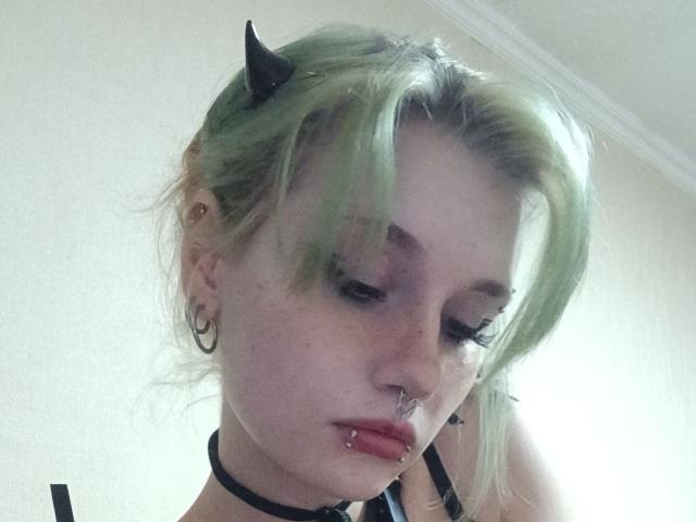 ShenaBatrum with green hair, black horns, nose ring, lip piercing, and hoop earrings, wearing a black choker, looking down with half-closed eyes, in a soft light.