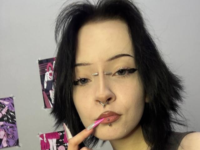 MarryGherardi with black hair, nose piercings, and lip piercing, touching her pink-lipsticked lower lip, wearing eyeliner, and looking at the camera.
