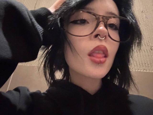 Marry Gherardi, met zwart haar, een bril, een neusring, een lip-piercing en rode lippenstift, draagt een zwarte hoodie en raakt haar hoofd aan. Borsten niet zichtbaar..