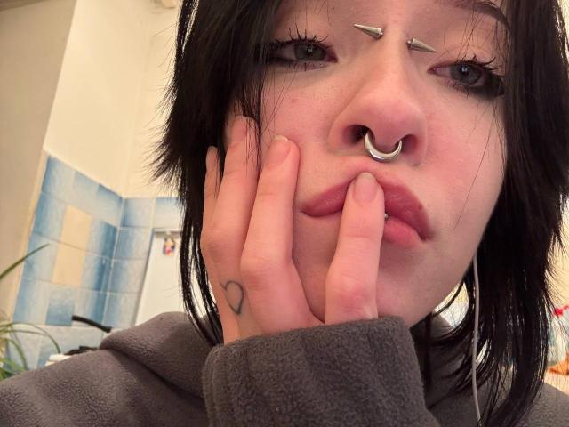 MarryGherardi with black hair, nose ring, and facial piercings, pouting with hand on cheek, wearing grey hoodie, visible heart tattoo on hand.