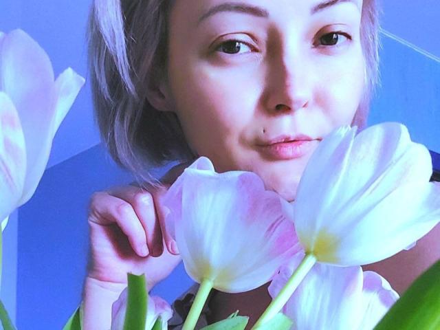 Lollyypop with pale pink hair, holding white tulips, looking at camera with subtle smile, blue light highlighting her face and petals. Her nose has a small black mole.