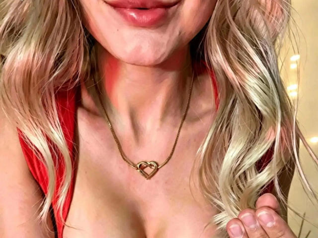 LaurenSommer-hot with blonde wavy hair, red top, and gold heart necklace, showing her tits and glossy pink lips, holding her hair on the right.