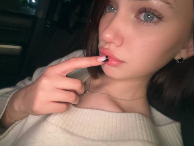 AlyceFujino with pale skin, blue eyes, and brown hair, touching her glossy pink lips with her finger, wearing a white ribbed top, and a thin necklace.