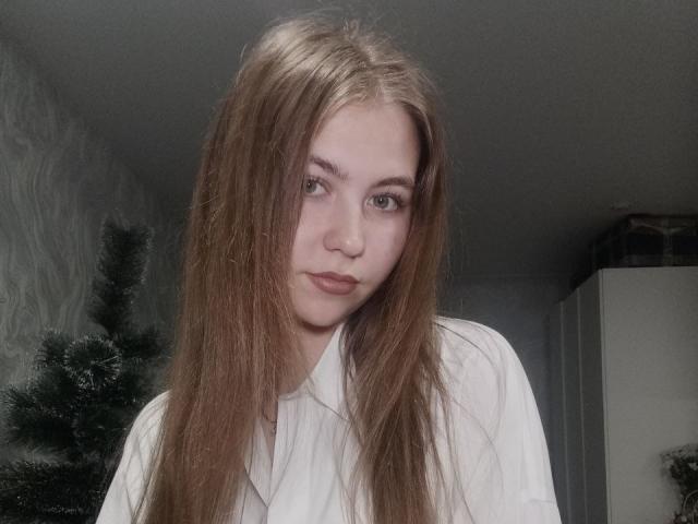 MargretWarriner has long, straight brown hair, pale skin, and green eyes. She's wearing a white shirt, looking slightly to the side with a subtle, serious expression.