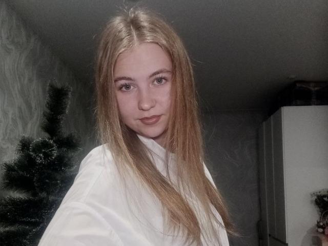 MargretWarriner with long blonde hair, wearing a white shirt, has blue eyes and a slight smile, standing next to a green Christmas tree in a dimly lit room.