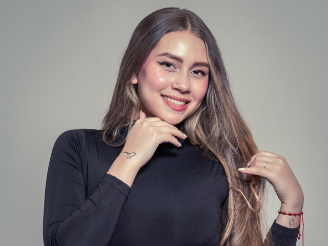 AlessiaCruz has long brown hair, pink eyeshadow, and a cute smile. She's wearing a black top, showing a small heart tattoo on her left hand. Boobs are not visible.
