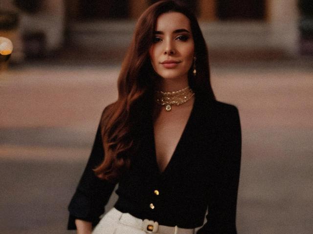 YukiCherry, com longos cabelos castanhos, blusa preta de decote em V, botão dourado, calças brancas, colar dourado e brincos, em pé, com uma aparência confiante, sexy e elegante..
