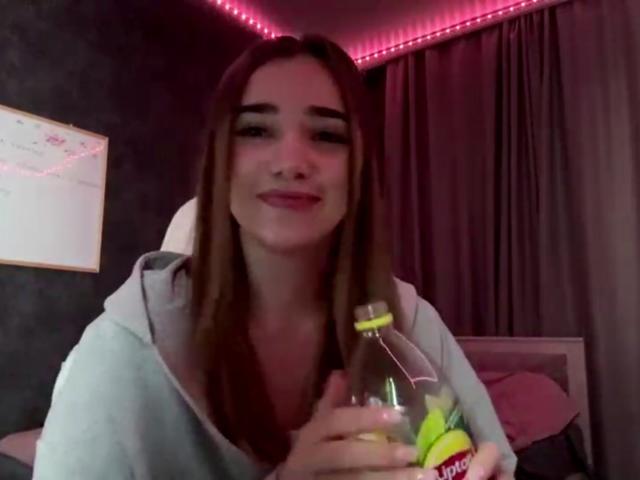 SandaScali mit langen, braunen Haaren, hellrosa Lippenstift, trägt eine graue Kapuzenjacke und hält eine durchsichtige Flasche mit einer Limette. Sie lächelt, im Hintergrund sind rosa LED-Streifen zu sehen..