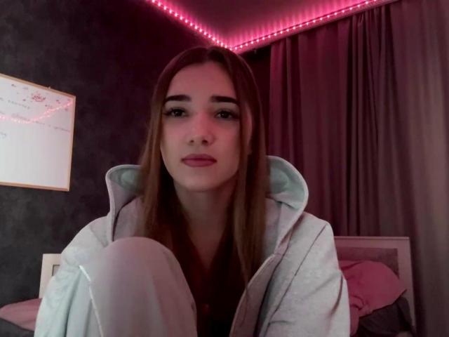 SandaScali with long brown hair, wearing a gray hoodie, sitting with knees up, looking at camera, pink LED strip lights in background, soft pink pillow, serious expression.