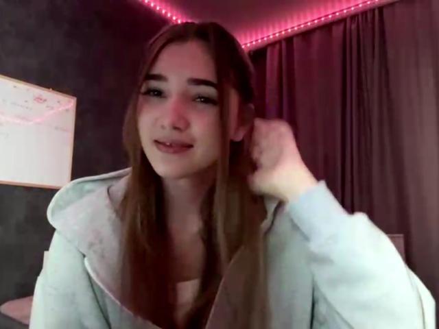 SandaScali with long brown hair, wearing a light grey hoodie, smiling slightly, hand in hair, pink LED strip lighting in background.