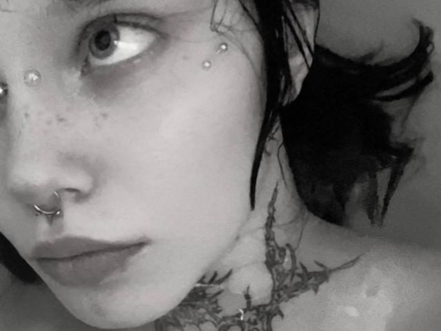 MariannaWarren's face in a black-and-white photo, showing a nose ring, freckles, eye piercings, and a detailed tattoo on her neck. She has wet, dark hair.