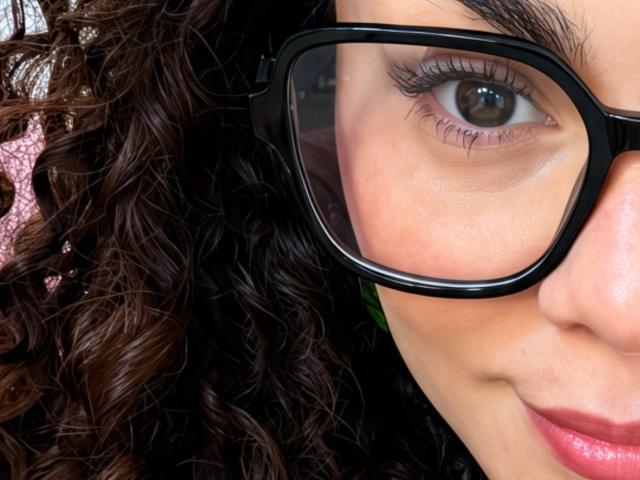 MissSunnyShine's face, showing curly brown hair, brown eyes with long lashes, and wearing black glasses, with a slight smile and pink lips, in a tight shot.