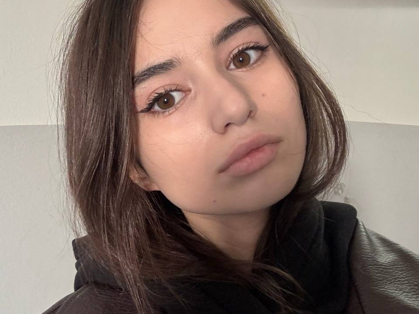 TonetteMatheu tem olhos castanhos, sobrancelhas grossas e cabelo castanho liso. Ela está vestindo um moletom preto, com uma expressão séria e os lábios levemente franzidos. Sua pele é clara e lisa..