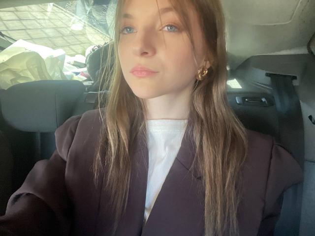 Annabell Morrall hat hellbraunes Haar, blaue Augen und rosafarbene Lippen. Sie trägt einen schwarzen Blazer über einem weißen Hemd und sitzt in einem Auto mit einem goldenen Ohrring..