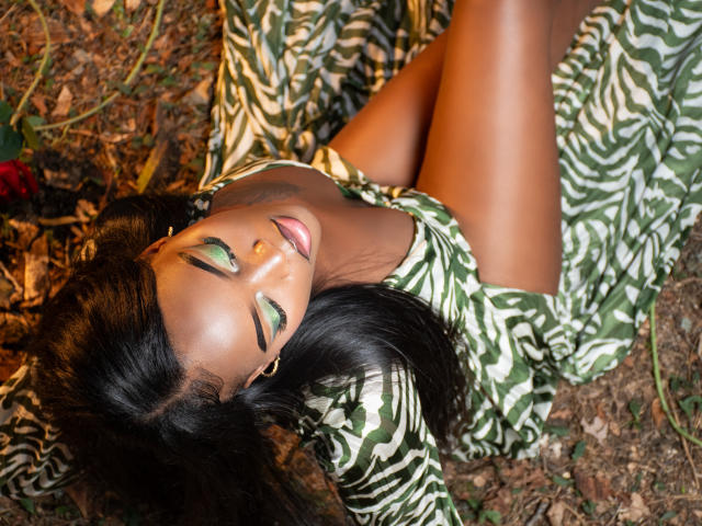 ZenyaEbony, avec sa peau foncée, son fard à paupières vert et ses cheveux noirs, est allongée sur le dos, vêtue d'une robe à rayures zébrées vertes et blanches, laissant apparaître ses jambes nues, et elle sourit..