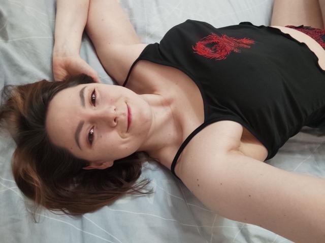 Patricia Casgrove liegt auf einem Bett, trägt ein schwarzes Tanktop mit einem roten Design und rote Spitzenunterwäsche. Sie hat braunes Haar, einen Nasenring und ein leichtes Lächeln..