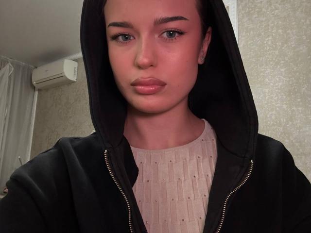 DrusillaMaiava with pinkish skin, black hoodie, and pink ribbed shirt, wearing earrings, has full lips, small nose, and looks straight into the camera. Her eyes are slightly downcast.