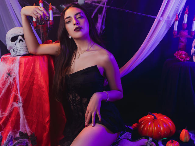 CelesteLarson, avec de longs cheveux bruns, du rouge à lèvres foncé, une robe noire sans bretelles, assise avec une jambe levée, un crâne sur la table, des bougies, un tissu rouge, des citrouilles, un éclairage violet et rouge..