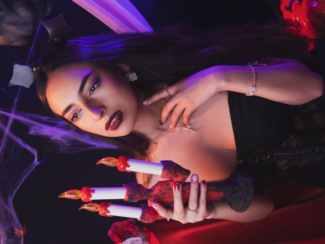 CelesteLarson met donkere lipstick, die rode en witte kaarsen vasthoudt, draagt een zwarte kanten jurk, een vleermuishoofdband en sieraden, en ziet er verleidelijk uit, met paars en rood licht..