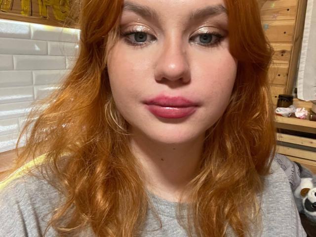 Marlyn Horbert hat rotes Haar, grüne Augen und rosafarbene, glänzende Lippen. Sie trägt ein graues Hemd und große Ohrringe. Ihre Haut ist hell mit feinen Sommersprossen..