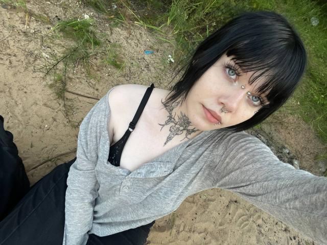 Marianna Warren, mit schwarzen Haaren, einem grauen Hemd, einem schwarzen BH und Tätowierungen, hat blaue Augen, eine Nasenring und einen Lippenring und sitzt auf sandigem Boden, während sie in die Kamera schaut..
