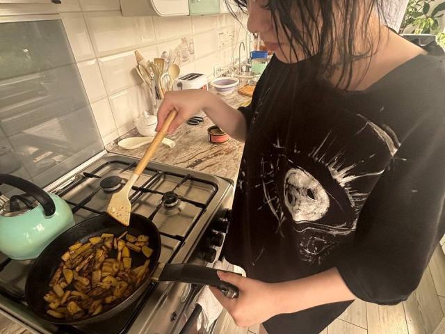 MariannaWarren with wet black hair, wearing a skull-print shirt, stirring a pan of caramelized onions on a stove with a wooden spoon, a teal kettle on the left.