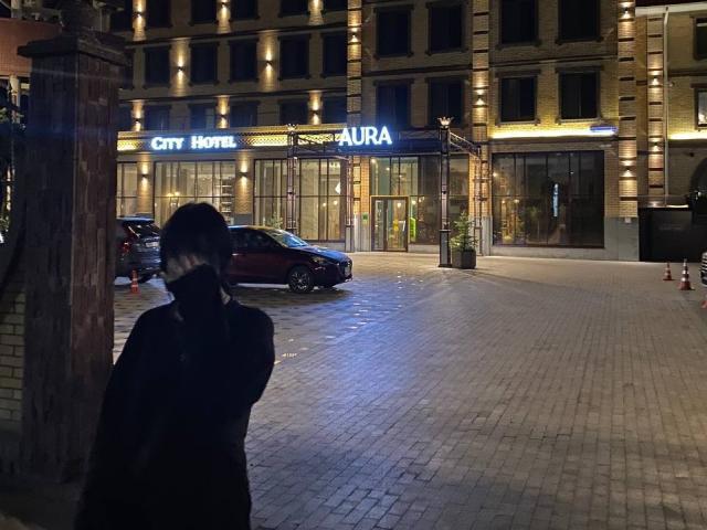 MariannaWarren stands with her back to the camera, wearing a dark coat, in front of a City Hotel Aura with a red car parked on the wet, cobblestone street.
