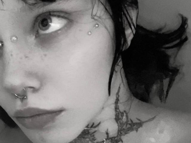 Grayscale selfie of MariannaWarren with dark hair, nose ring, freckles, tattoos on neck, and piercings on forehead, looking to the side with serious expression. Her tits are visible.