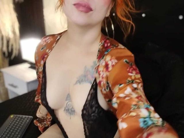 LilithSnop, avec ses cheveux orange, portant une robe à fleurs, de la lingerie en dentelle noire, montrant ses seins et ses tatouages, assise à un bureau avec un clavier. Rouge à lèvres rouge. Boucles d'oreilles cerceaux..
