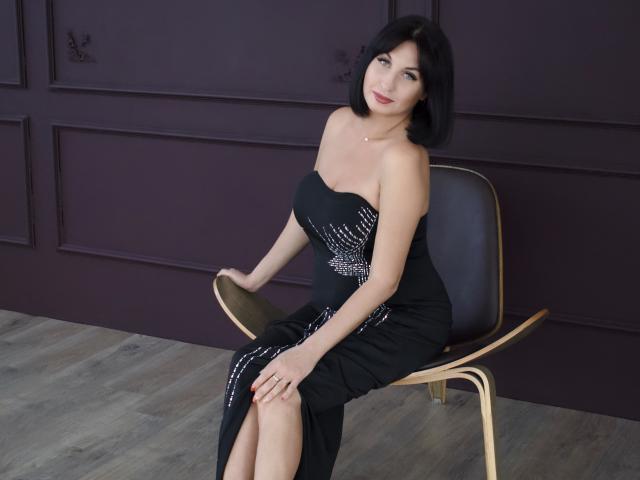 OlympiaKassel com cabelo curto preto, batom vermelho, vestido preto sem alças, detalhes prateados brilhantes, sentada em uma cadeira de madeira, mãos nos joelhos, chão de madeira, parede roxa, olhando para a câmera, sorrindo..