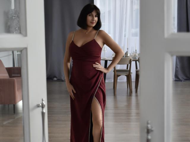 OlympiaKassel, com um corte de cabelo curto e escuro, vestindo um vestido vermelho com uma fenda alta, mostrando sua coxa nua, com as mãos nos quadris, em pé em um quarto com portas brancas..