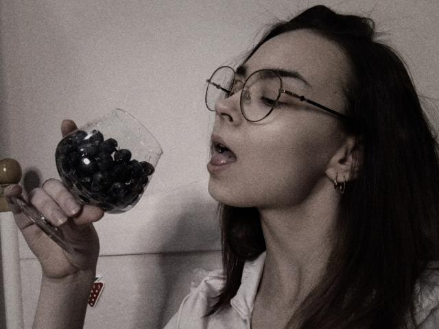 LelaVivo with glasses, tongue out, holding a glass of blueberries, white shirt, dark hair, and small earrings, in a dark, grainy photo.