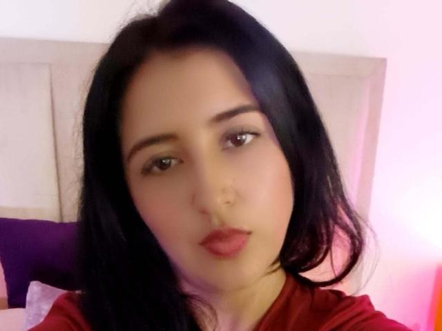 NicollJeans has black hair, brown eyes, and red lips, wearing a red shirt, with a slight pout, looking directly at the camera in a soft-lit room.