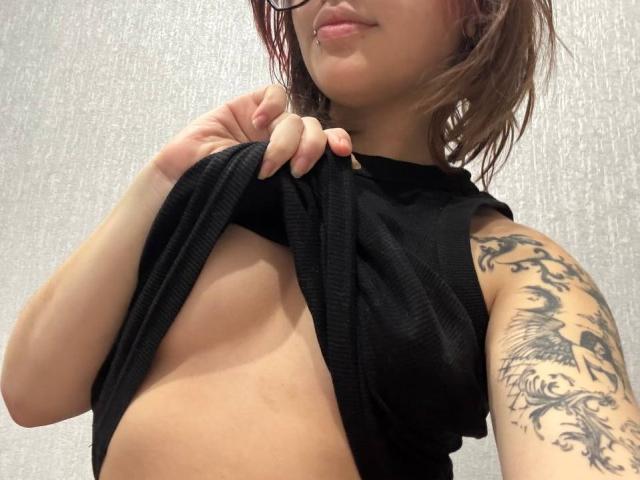 JackelynGile alza una maglietta nera per mostrare il suo seno nudo, con un tatuaggio sul braccio destro, indossa degli occhiali e ha un leggero sorriso. I suoi capezzoli sono visibili..