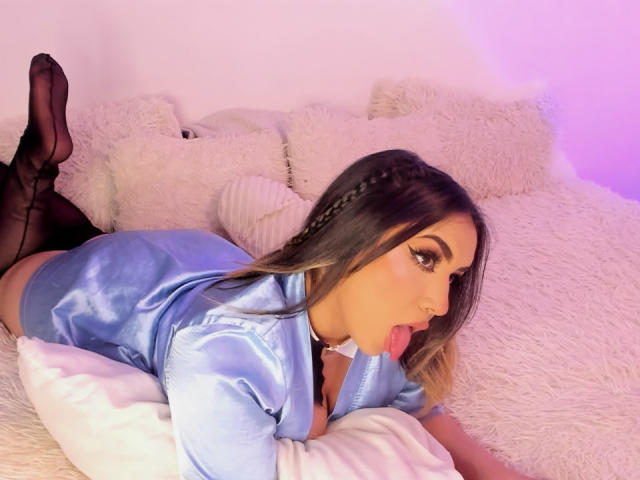 MelissaLust lying on a bed, wearing a shiny blue satin shirt, black thigh-high stockings, and licking a pillow, with her tongue out, looking sexy and cute.