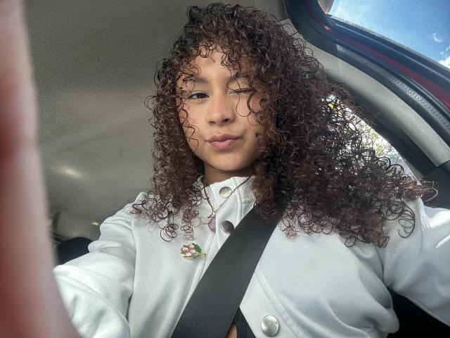 Sofia, passionnée, avec des cheveux bruns bouclés, portant une chemise blanche, une ceinture de sécurité autour de la poitrine, une épingle à fleur rose, et une expression boudeuse, assise dans une voiture. La lumière du soleil illumine son visage..