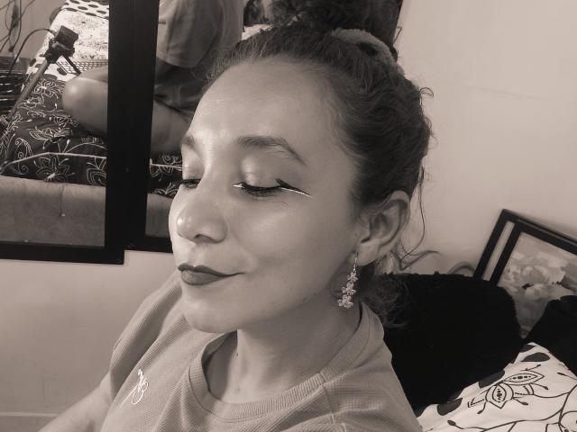 AmberDemons with eyes closed, wearing makeup, has glossy skin, dark lipstick, and long eyeliner, smiling slightly, wearing a tee, and floral earrings, in a black-and-white photo.