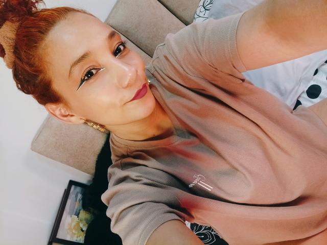AmberDemons with red hair in a bun, wearing a pinkish-brown shirt, showing a smile, eyeliner, gold earrings, and holding a selfie. Boobs partially visible, nipples not shown.
