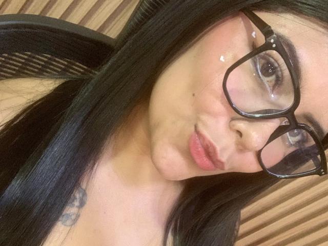 CatalinaReiyna with long black hair, pouty pink lips, and big black glasses, pucker-lips selfie, subtle tattoos on her neck, fair skin, and a cute small mole on her cheek.