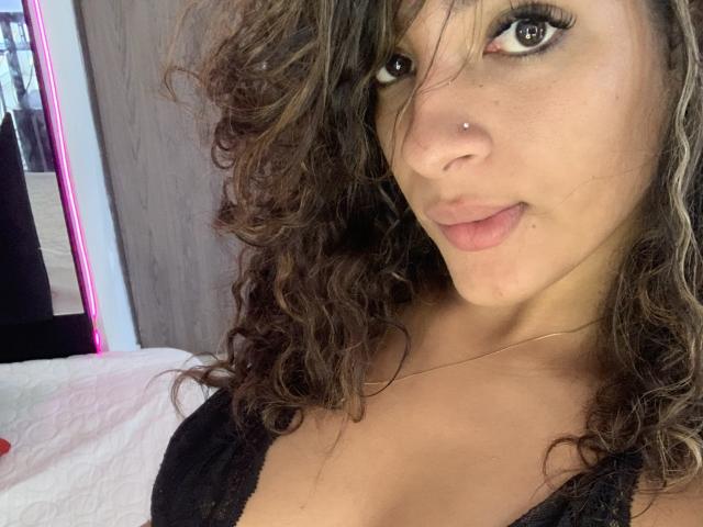 A CristalAndRichard tem cabelo castanho encaracolado, um piercing no nariz e está vestindo lingerie de renda preta, mostrando seus seios e um leve sorriso. Seus olhos estão olhando para a câmera..