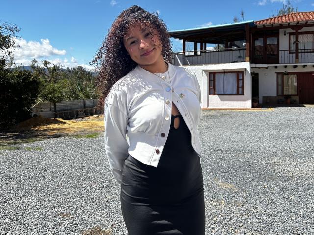 SofiaPassionne con i capelli castano ricci, giacca bianca, vestito nero, mani dietro la schiena, in piedi su ghiaia, sorridente, cielo azzurro limpido, casa bianca e marrone sullo sfondo..