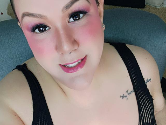 GiiannaMorrys avec fard à paupières rose, blush rose, piercing au nez, débardeur noir, tatouage sur l'épaule droite, souriant vers l'appareil photo, montrant ses seins. Peau claire, cheveux bruns, rouge à lèvres rose..