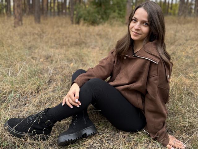 SaraUili with brown jacket, black ribbed pants, and black boots, sitting on grass, smiling, brown hair, forest background, hands on knee, slightly leaning back.