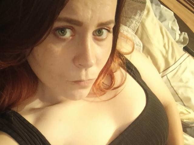 FannyNorah, con la pelle chiara, gli occhi verdi e i capelli rosso-marrone, indossa una canotta nera, guarda in alto con un'espressione leggermente confusa, mostrando piccoli seni e la clavicola..