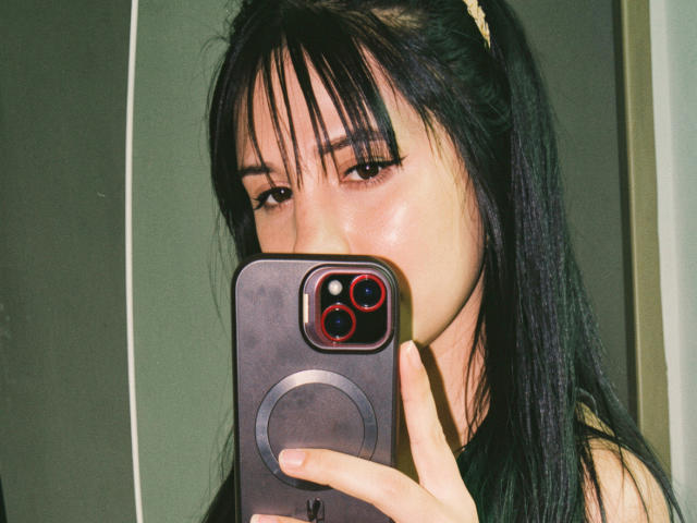 Kkatalina mit dunklem, glattem Haar und einem blonden Haarreif, die ein Selfie mit einem roten und schwarzen Telefon macht, ihr Gesicht ist leicht glänzend, und man sieht ihre Nase und Augen..
