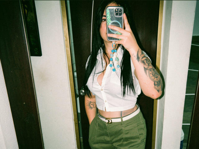 Kkatalina, con cabello negro largo, vestida con una camisa blanca de botones que deja ver sus pechos, pantalones verdes y un cinturón blanco, tomando una selfie frente al espejo con tatuajes en los brazos..