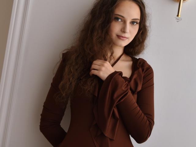 MilenaSparkle has long, curly brown hair and wears a brown, long-sleeve dress with a ruffled neckline, standing against a white wall, smiling slightly with a gentle hand on her chest.