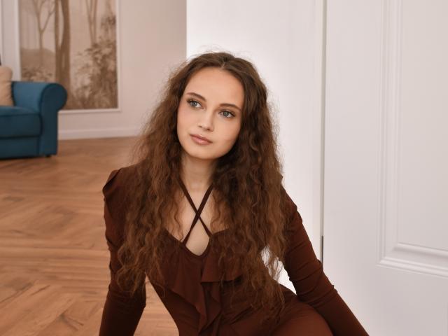 MilenaSparkle has curly brown hair, green eyes, and is wearing a brown dress with a crisscross neckline, sitting on a wooden floor, smiling slightly.