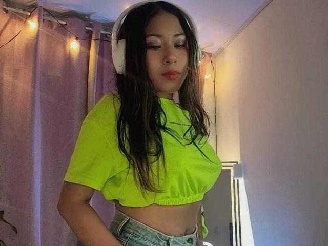 SiaraBonny, con cabello largo y oscuro, vistiendo una camiseta corta de color verde neón, audífonos blancos y jeans azules, mostrando sus pequeños senos y su trasero ligeramente curvado..